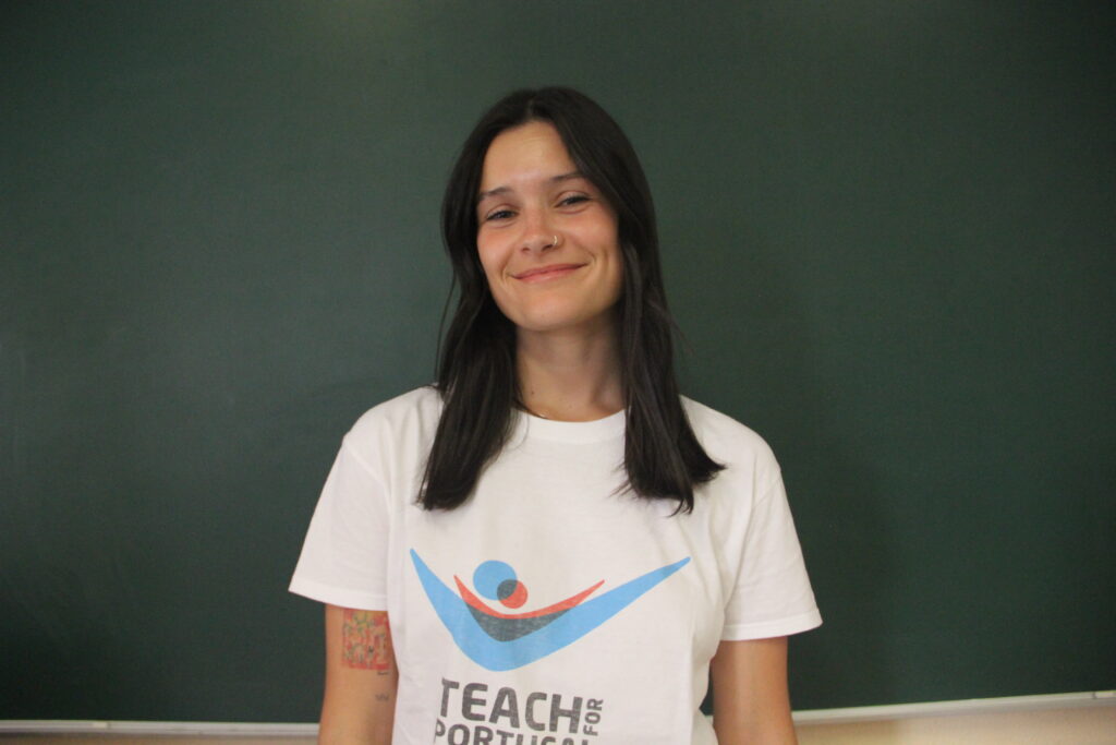 Maria Matos, Mentora 5ª Geração Teach For Portugal a usar a camisola com o logótipo da Teach For Portugal com um quadro de ardósia como fundo