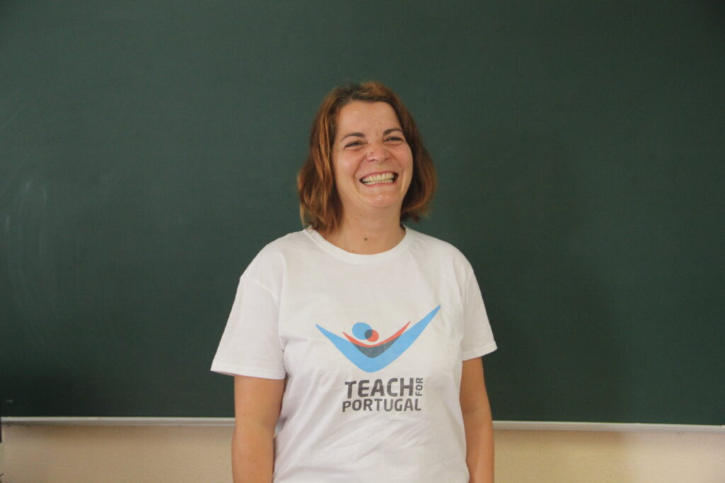 Alexandra Barreiros, Mentora 5ª Geração Teach For Portugal com um sorriso enorme a usar a camisola com o logótipo da Teach For Portugal com um quadro de ardósia como fundo