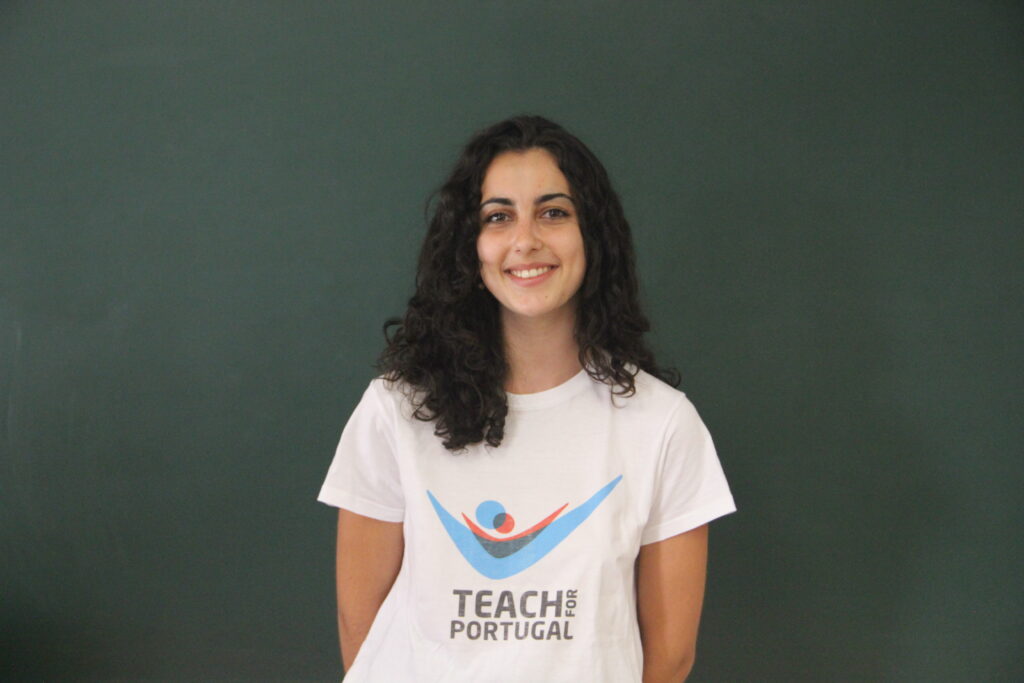 Ângela Matias, Mentora 5ª Geração Teach For Portugal a usar a camisola com o logótipo da Teach For Portugal com um quadro de ardósia como fundo