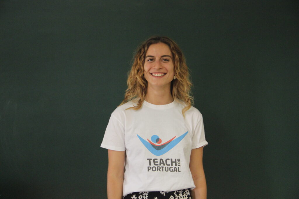 Sara Rodrigues, Mentora 5ª Geração Teach For Portugal a usar a camisola com o logótipo da Teach For Portugal com um quadro de ardósia como fundo